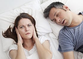 Woman covering ears next to snoring man in bed